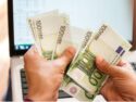 Plik banknotów euro w dłoni, w tle ekran komputera